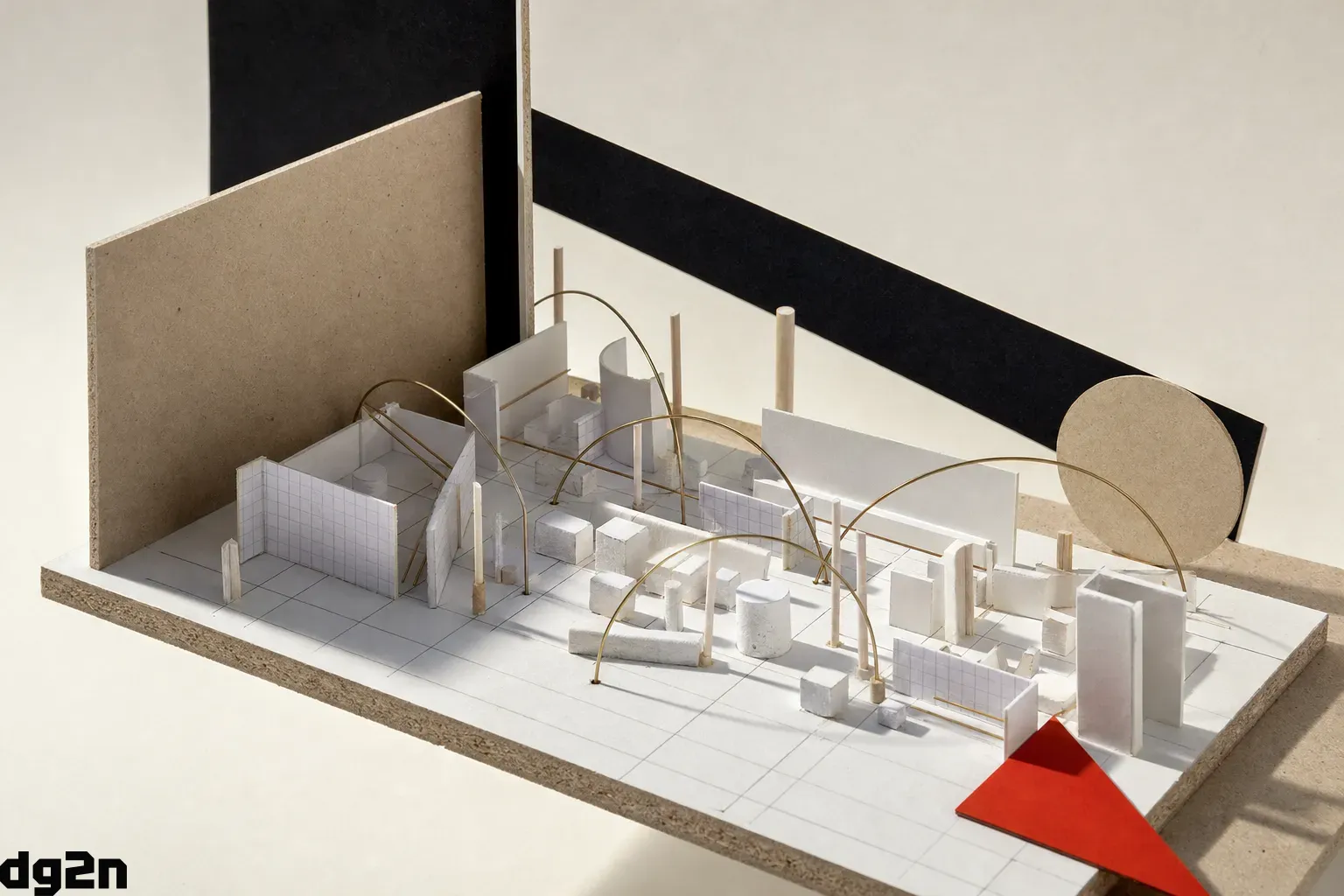 A physical study model of a miniature retail layout built from grid paper, white cardstock, plaster blocks, and brass wire arcs tracing pathways over modular shelf partitions.